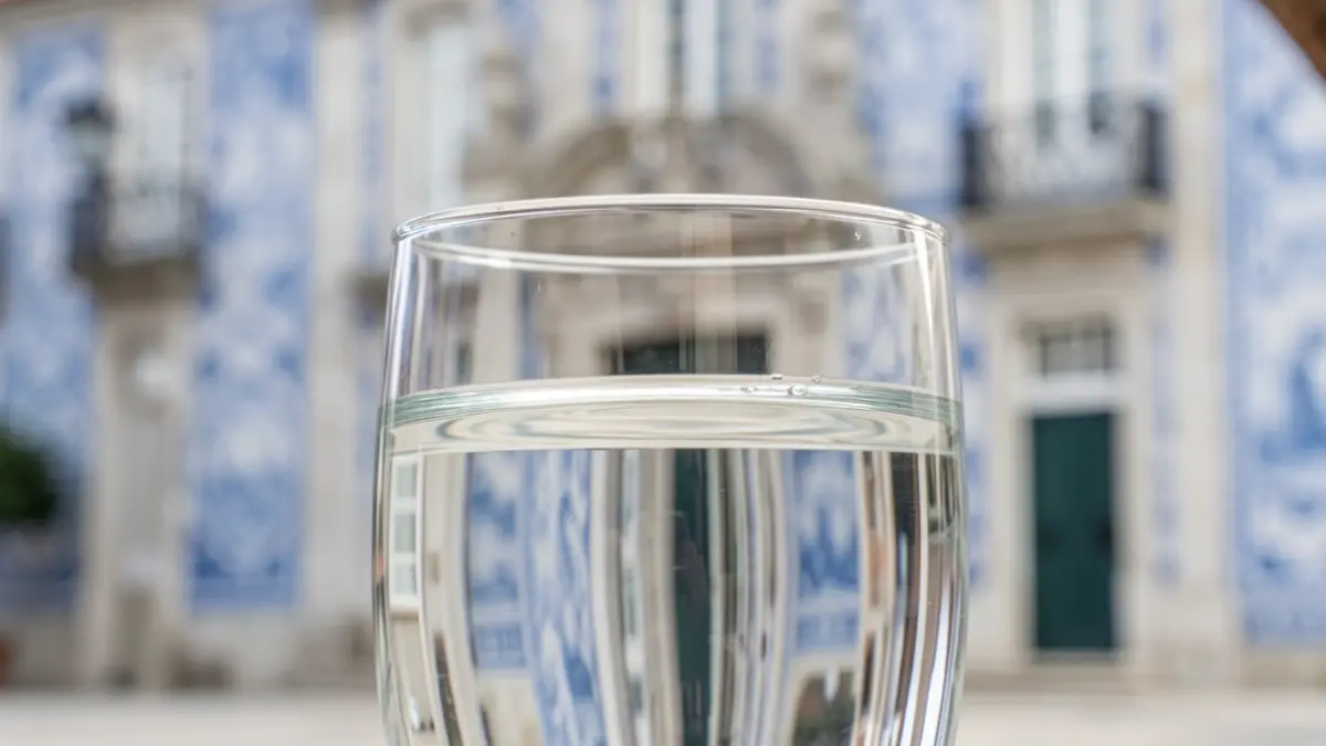 Imagem genérica de um copo de água límpida com reflexos, em frente a uma fachada de edifício público com azulejos portugueses.