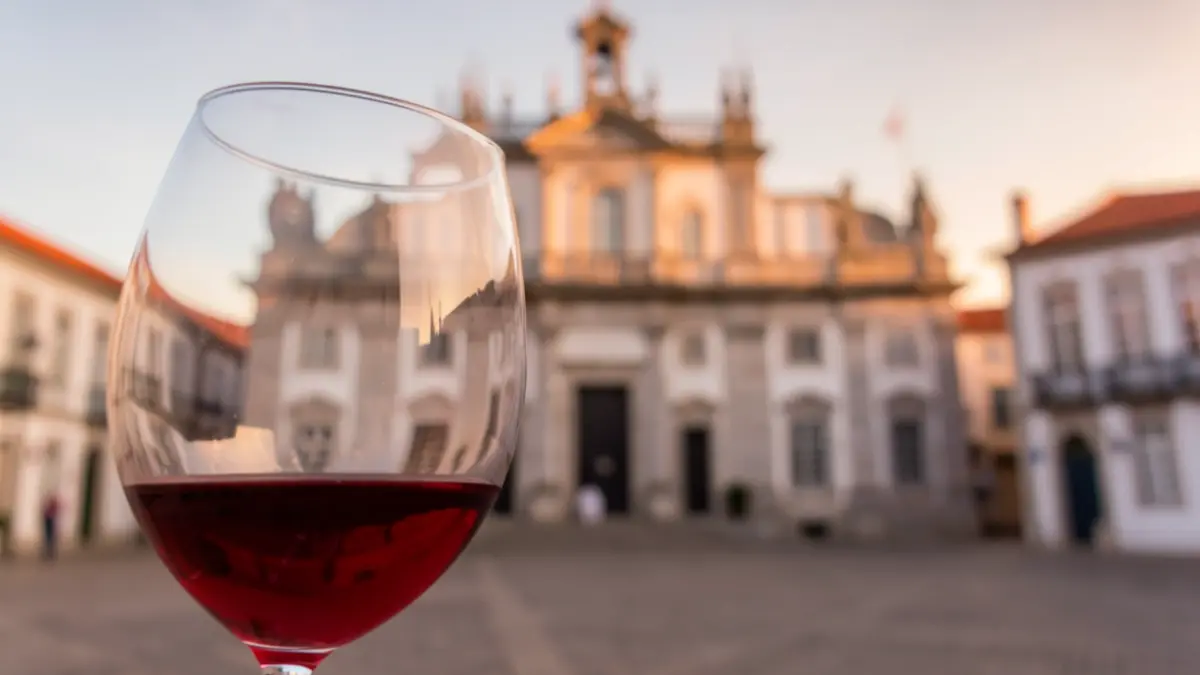 Imagen de una copa de vino tinto en primer plano, con la fachada de un edificio municipal portugués desenfocada al fondo.